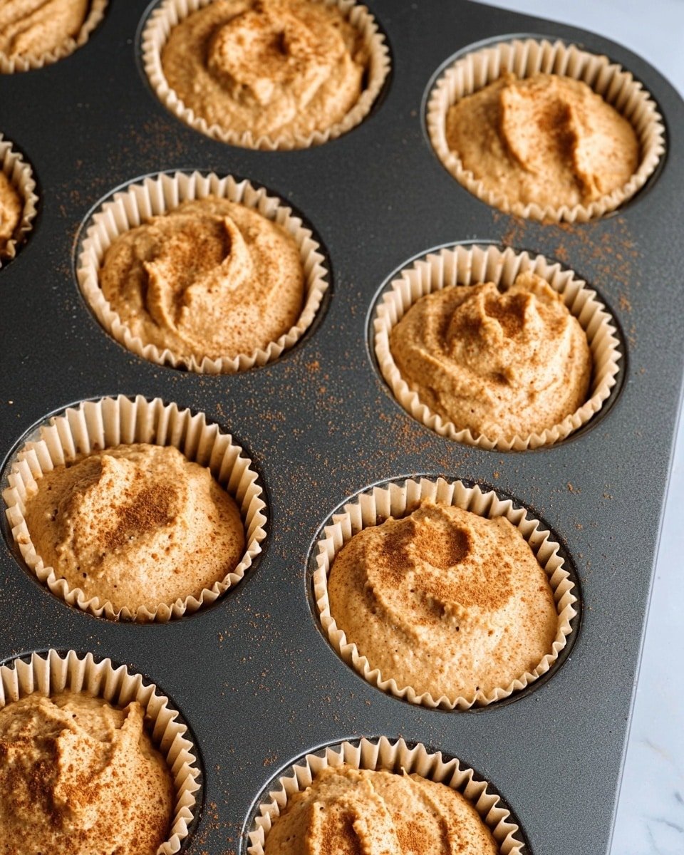 The image shows a dark metal muffin tray filled with eleven raw cupcake batters, each in beige paper liners. The batter is light brown with specks of darker ingredients, giving a slightly rough texture. Each muffin cup is about three-quarters full, with the batter unevenly spread and showing soft peaks and swirls. There is a light sprinkling of a finer brown powder, likely cinnamon or nutmeg, on the tops of the batter. The background where the tray is placed is a white marbled texture. Photo taken with an iphone --ar 4:5 --v 7